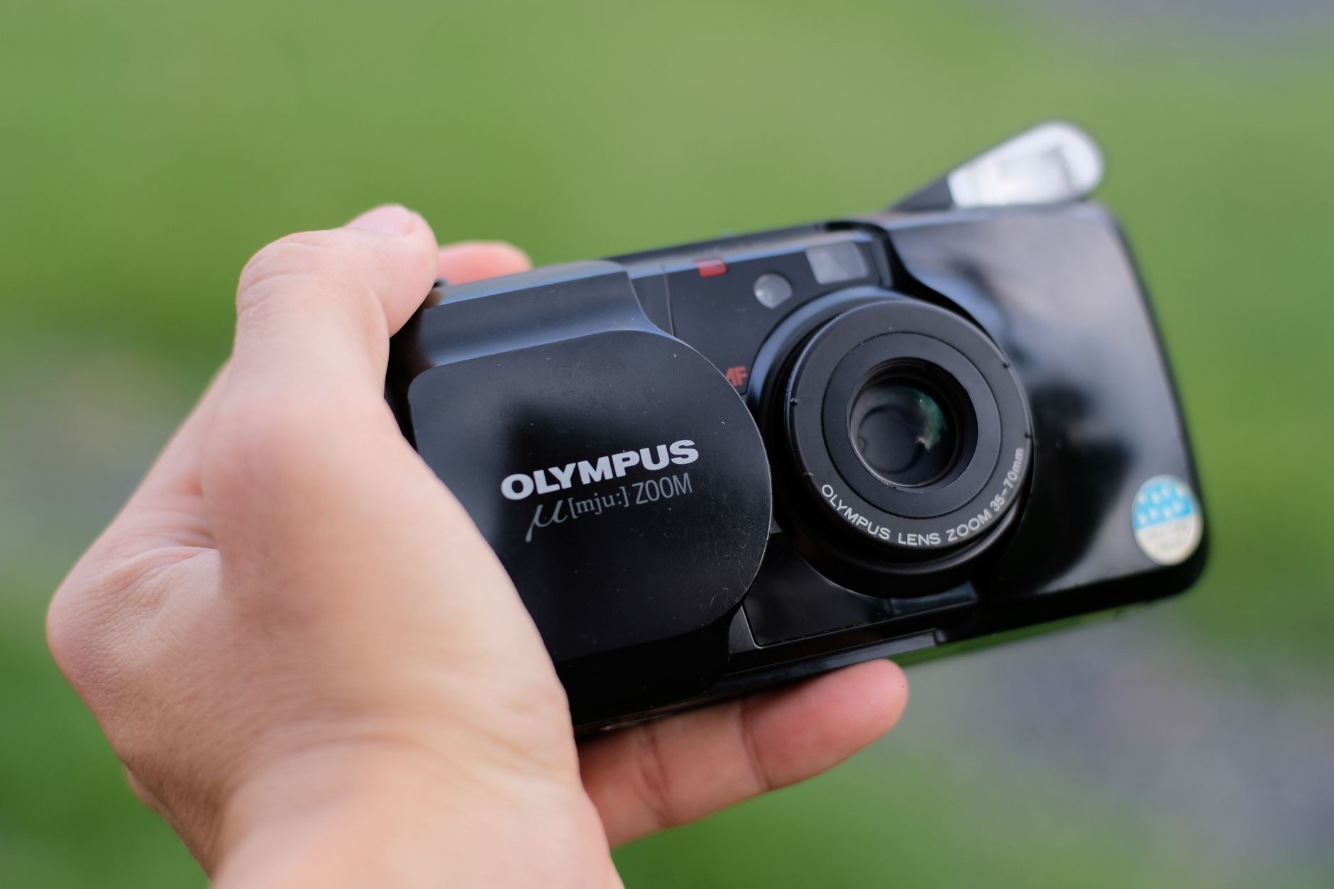 Close-up of a hand holding a vintage Olympus camera outdoors with a blurred green background.