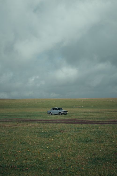 Free A solitary classic car parked in a wide open field beneath an overcast sky, exuding a mysterious and timeless atmosphere. Stock Photo