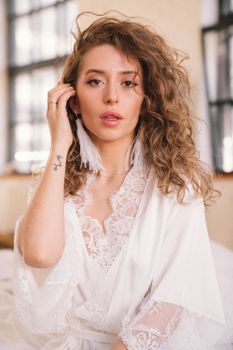 Portrait of a young woman in a white lace robe with wavy hair and elegant accessories, indoors.