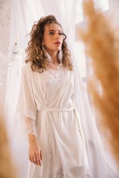 A young woman elegantly dressed in a white lace robe stands indoors with soft natural lighting.