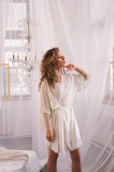 A woman in a white robe poses elegantly in a bright, luxurious setting with a chandelier.