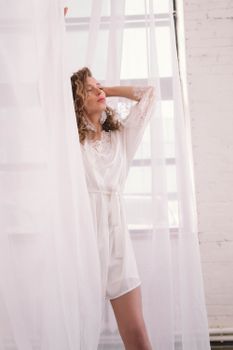 A serene portrait of a woman in white attire posing behind sheer curtains indoors.