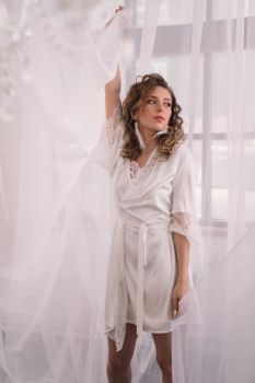 A woman in a white robe poses gracefully by sheer curtains in a softly lit room.