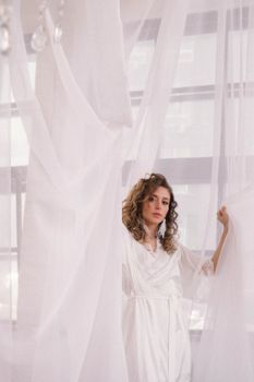 A beautiful bride stands in a white robe, surrounded by flowing drapes, creating a serene atmosphere.