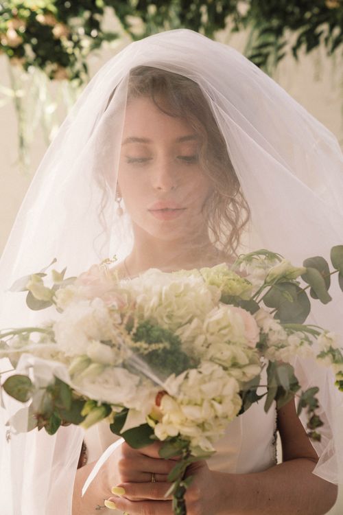 Free A serene bride holding a white and green bouquet, elegantly draped in a veil, outdoors. Stock Photo