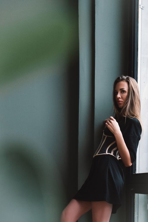 Free A woman in a black dress standing by a large window, exuding elegance and poise. Stock Photo
