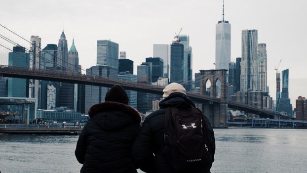 Two people enjoy the iconic view of the Manhattan skyline and Brooklyn Bridge from the riverbank.