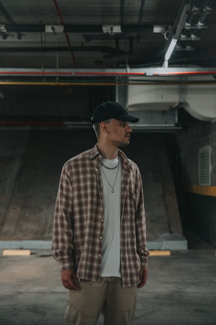Stylish young man in checkered shirt and cap standing in dimly lit urban setting.