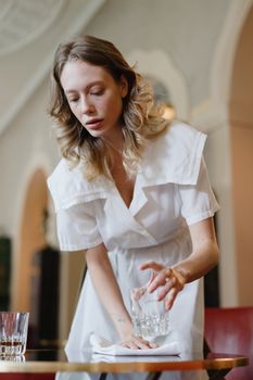 Graceful woman in a white dress setting a table with a glass indoors, capturing elegance and poise.