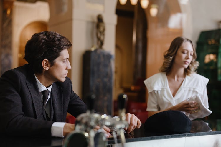 Young Man In A Suit Sitting At The Bar Counter Watching The Waitress Wiping Glass