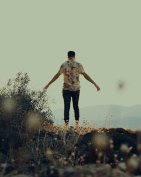 A man stands on rocky ground surrounded by nature, gazing at the distant landscape.