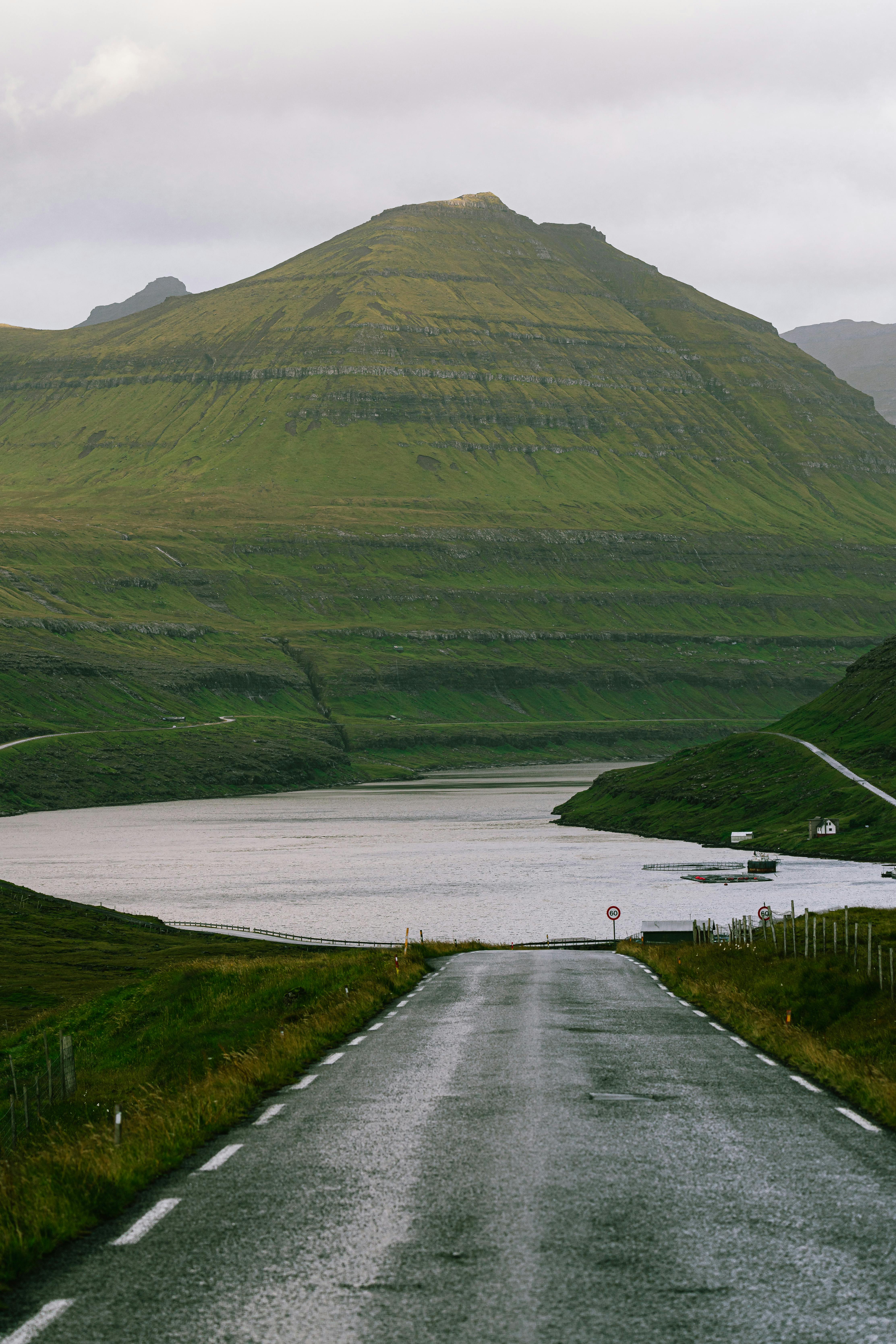 Paved road extending towards a tranquil lake and green mountain under a cloudy sky.