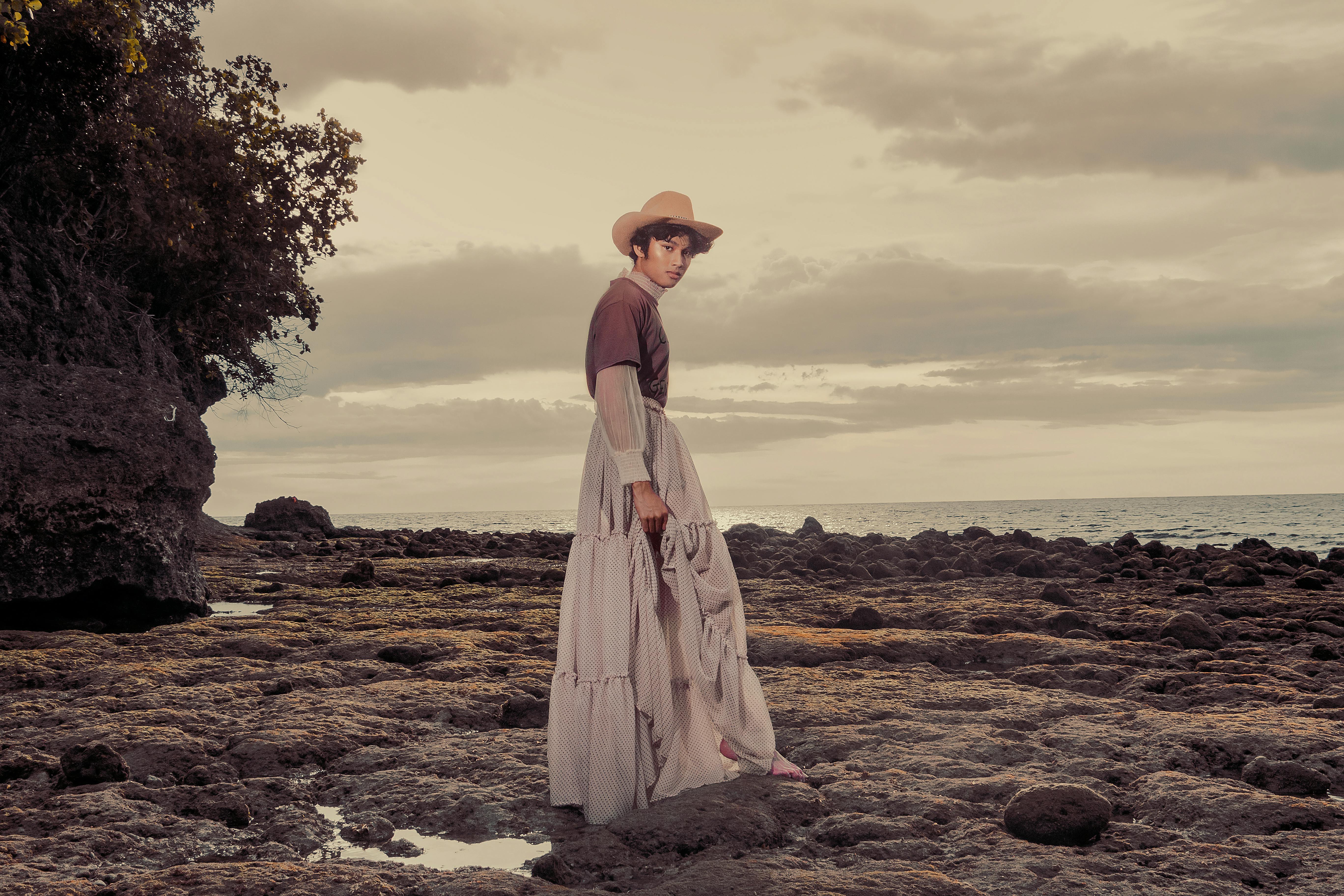 穿着时尚服装的雌雄同体模特在日落时在岩石海滩上时尚地摆姿势