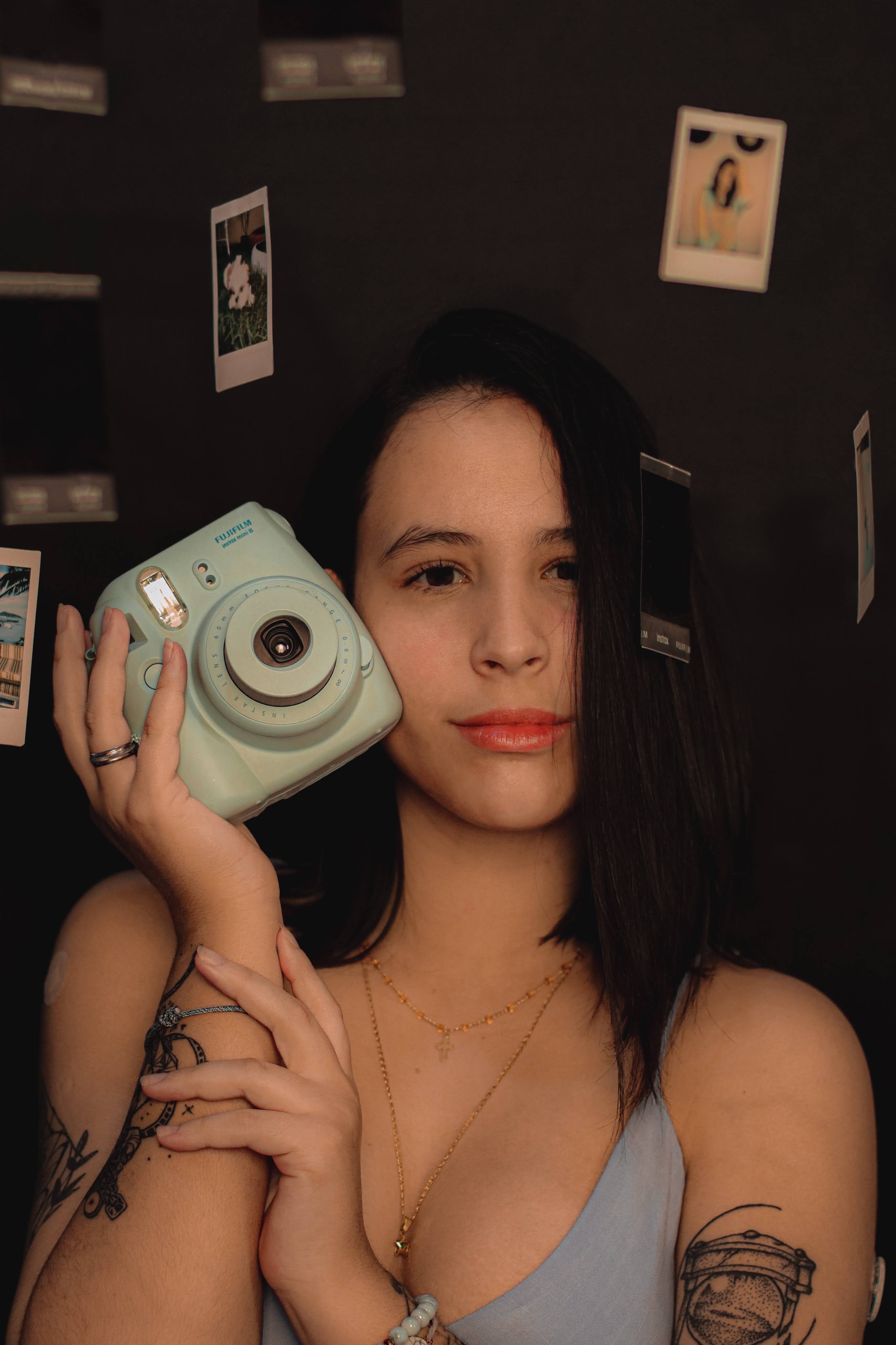 Caucasian woman holding a teal polaroid camera with floating photos around her.