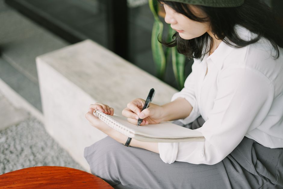 白いブラウスを着た若い女性が屋外でノートにメモを取っている。