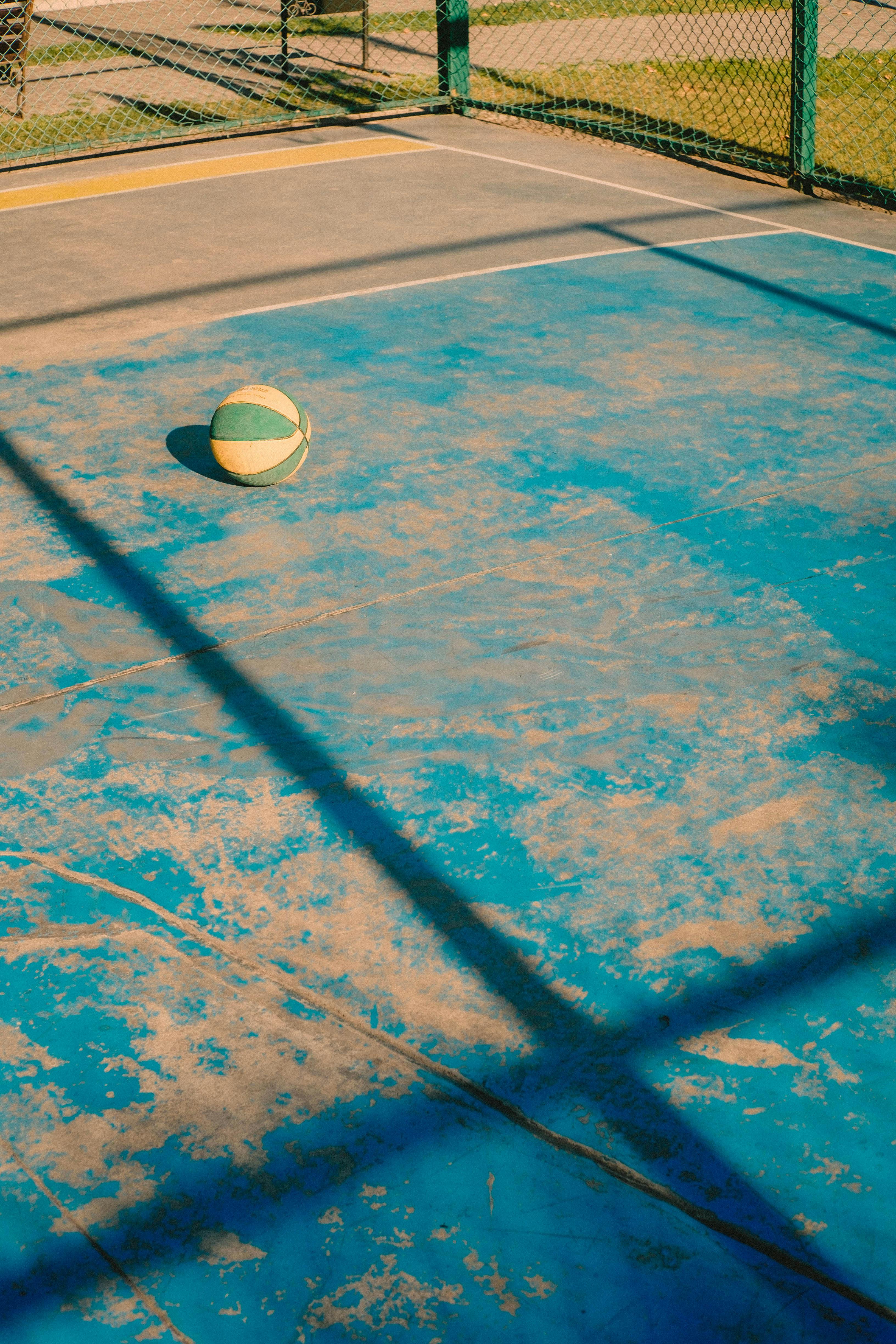 A single basketball on a sunlit, worn-out outdoor court with shadows.