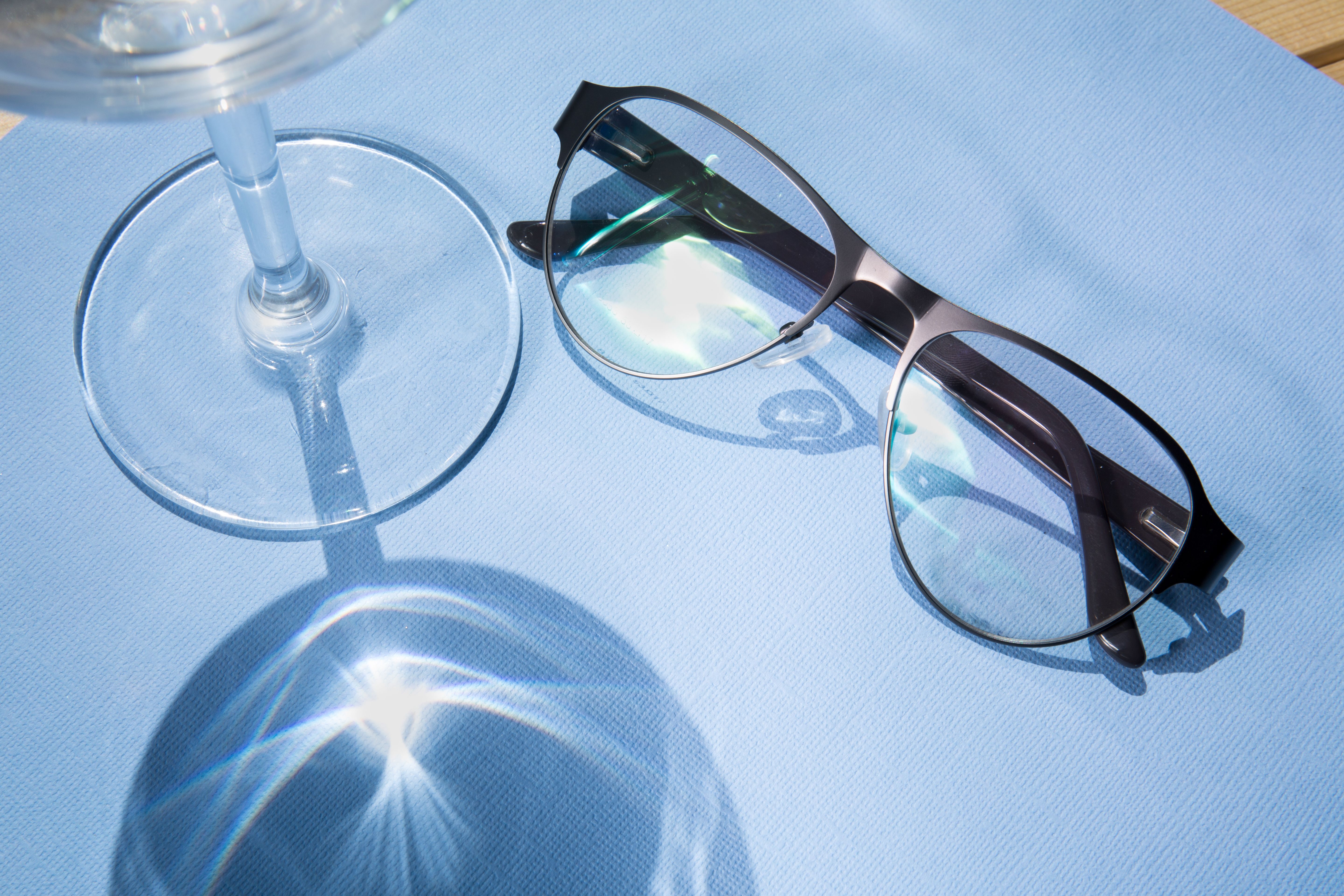 Stylish eyeglasses and wine glass on a blue textured surface, highlighting reflections and shadows.