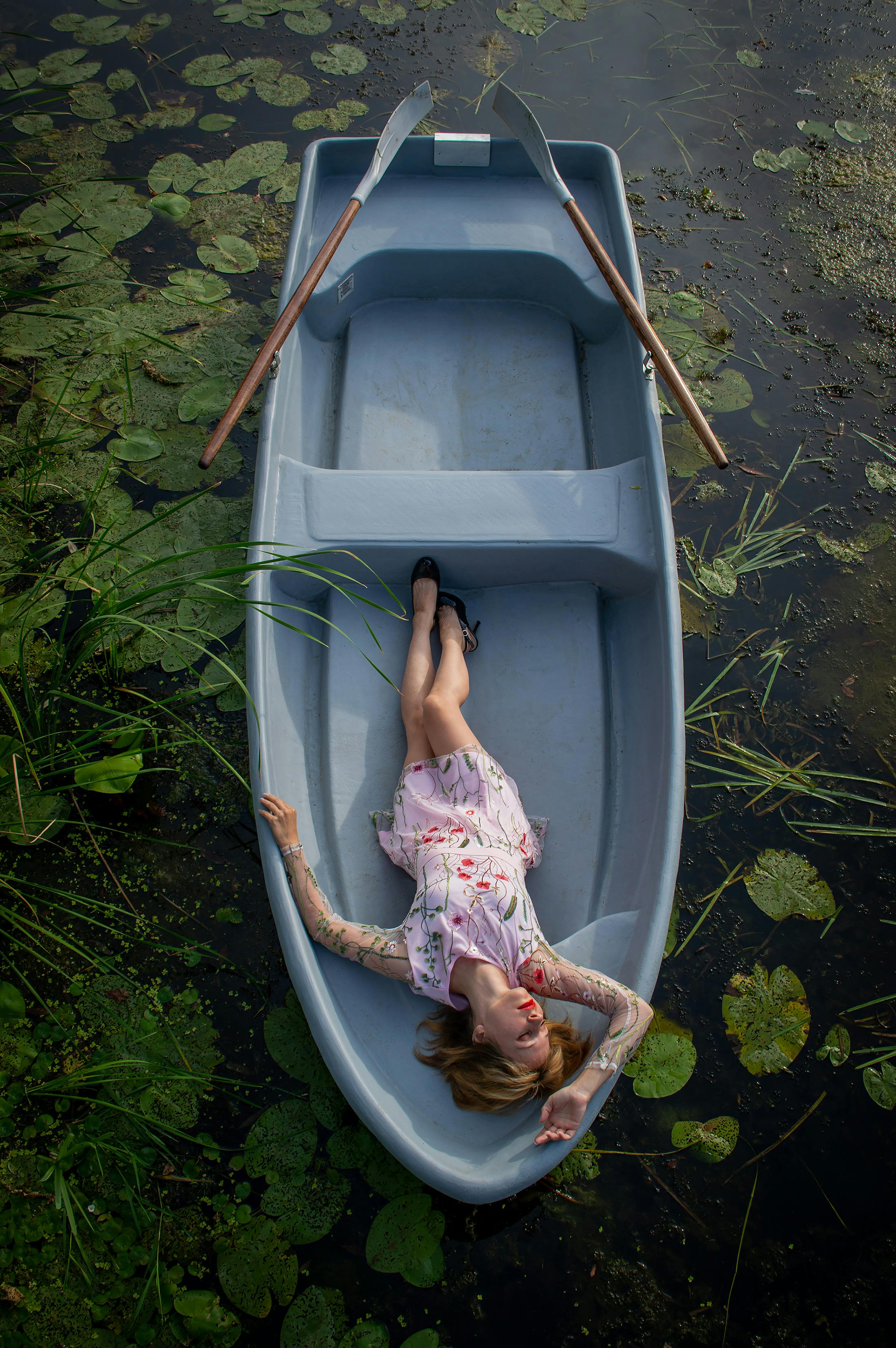 Aerial view of a woman in a floral dress lying in a boat on a pond with lilypads.