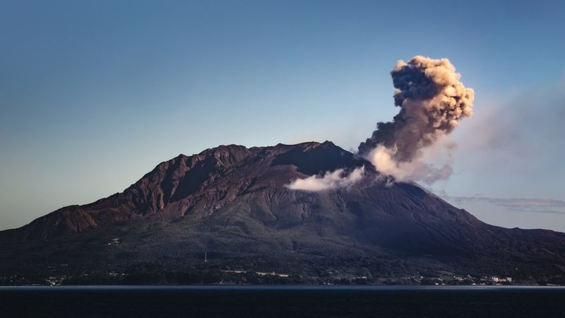 Sakurajima volcano actively erupting with ash plume rising against blue sky, viewed across Kinko Bay
