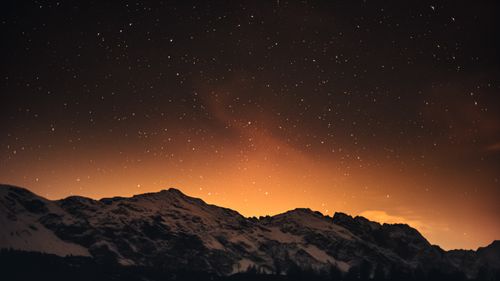 Free Captivating view of starry night sky over snow-covered mountains in Italy with a warm, orange glow. Stock Photo