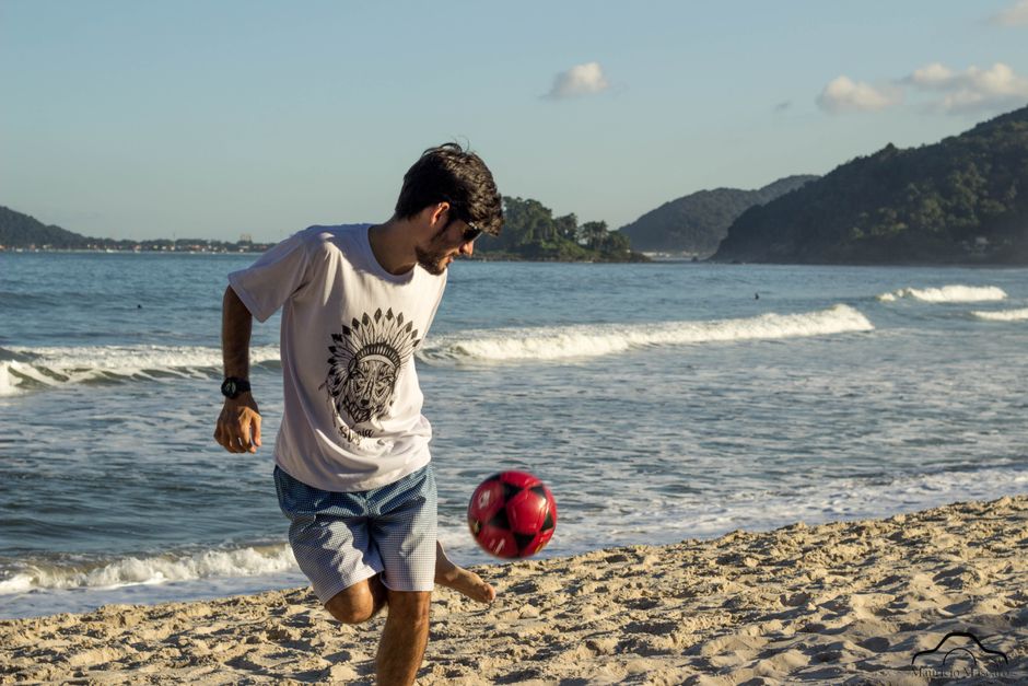 Young man enjoying playing soccer on a sunny beach with waves and mountains in the background.