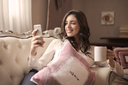 Mulher Em Camisa Branca De Mangas Compridas Segurando Um Smartphone Sentada Em Um Sofá Tufado