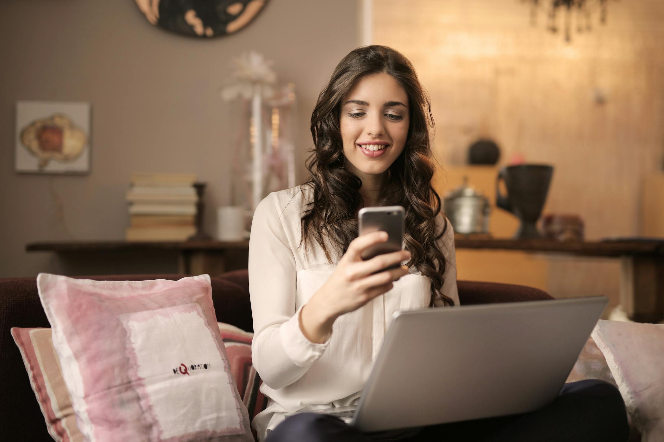 young female sitting on couch looking at phone with laptop in front of her
