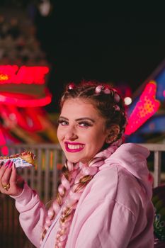 A smiling young woman with braided hair and vibrant makeup enjoying food at a night fair.