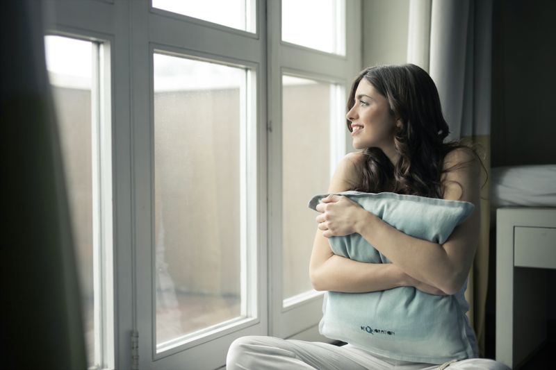 A woman sitting by a sunny window, hugging a cushion and smiling softly