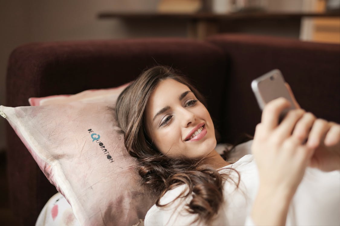 Free Woman in White Top Holding Smartphone Lying on Couch Stock Photo