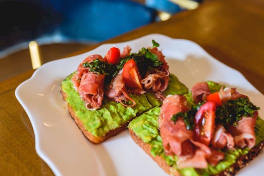 Delicious avocado toast topped with prosciutto, cherry tomatoes, and herbs on a white plate.