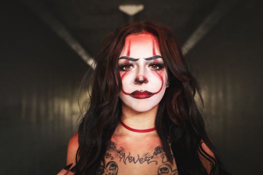 A dramatic portrait of a woman with clown makeup and tattooed chest, captured indoors.
