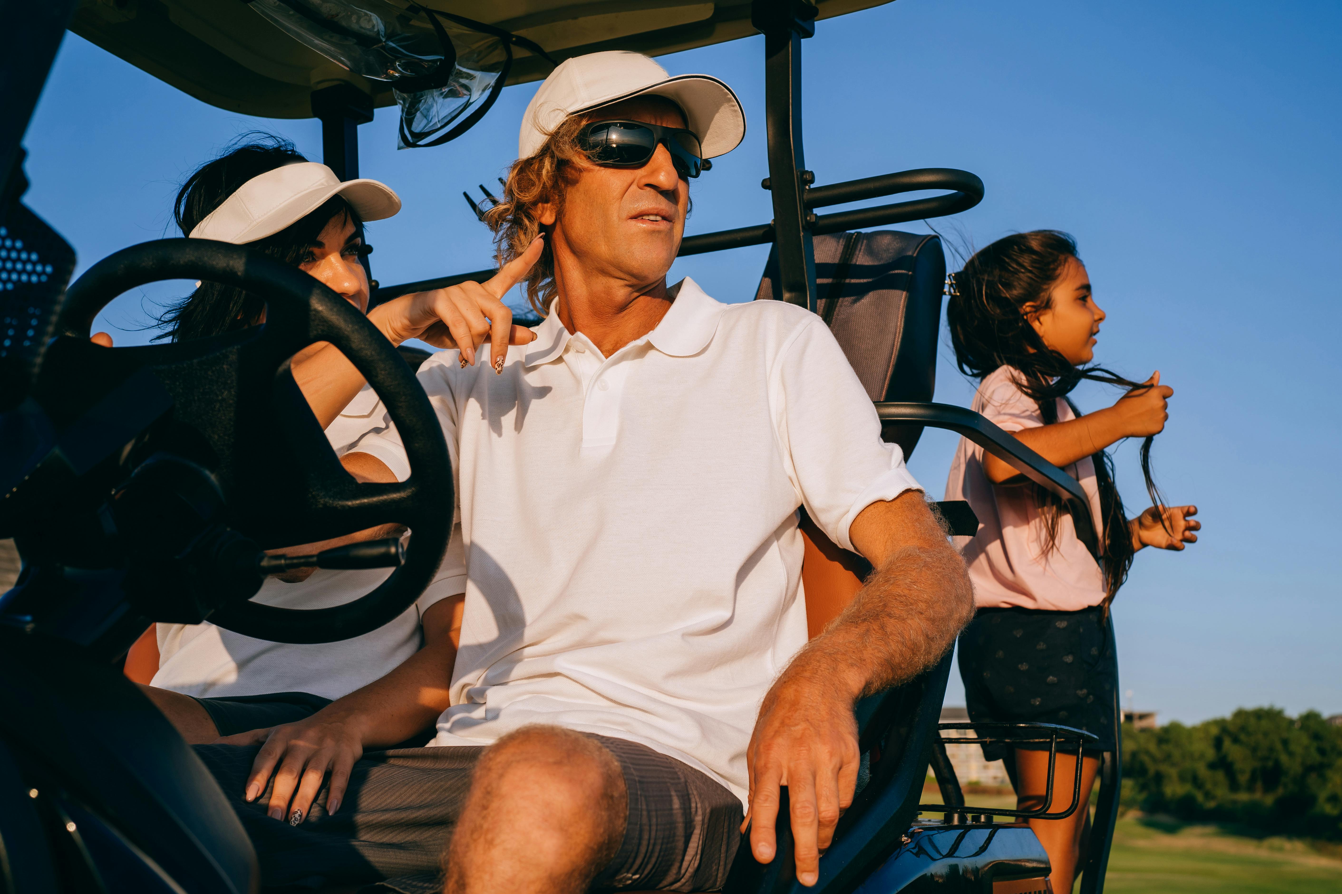 a man in white polo shirt and hat riding a golf cart with woman and a girl