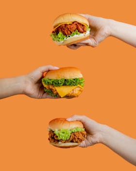 Three chicken burgers with lettuce and cheese, held in hands against a vibrant orange background.