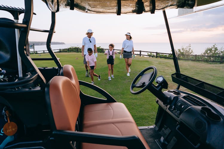 Happy Family Walking On Green Grass Field Near Golf Cart