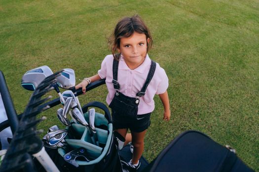 A child in casual attire stands beside a golf cart, holding a golf club outdoors on a green lawn.