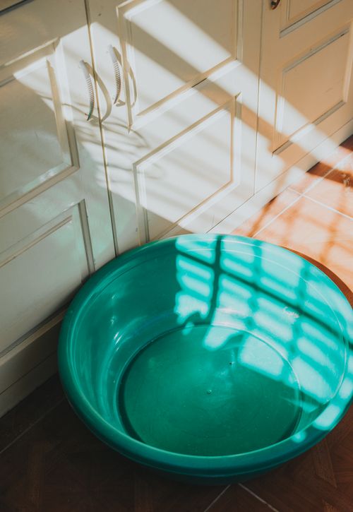 Free A turquoise plastic basin placed in a sunny kitchen corner, casting shadow patterns on surrounding cabinets. Stock Photo