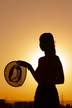 Silhouette of a woman holding a hat against a vibrant sunset sky.