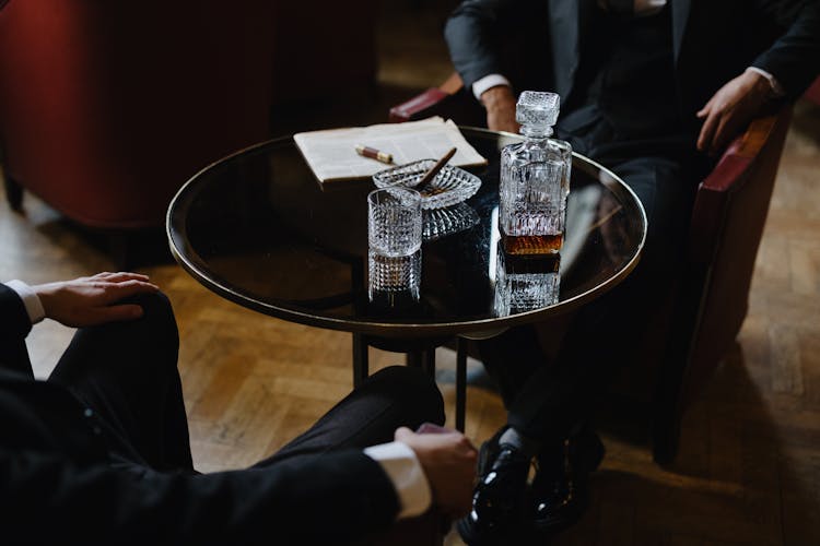 Men In Black Suits Sitting On Armchairs Beside Round Table With Pitcher And Glass