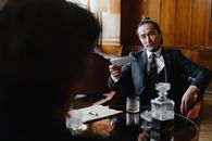 Man in Black Suit Sitting Beside Table and Holding a Paper