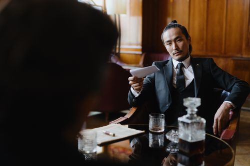Free Asian man in a black suit sitting at a table holding a paper in a formal, retro-style setting. Stock Photo