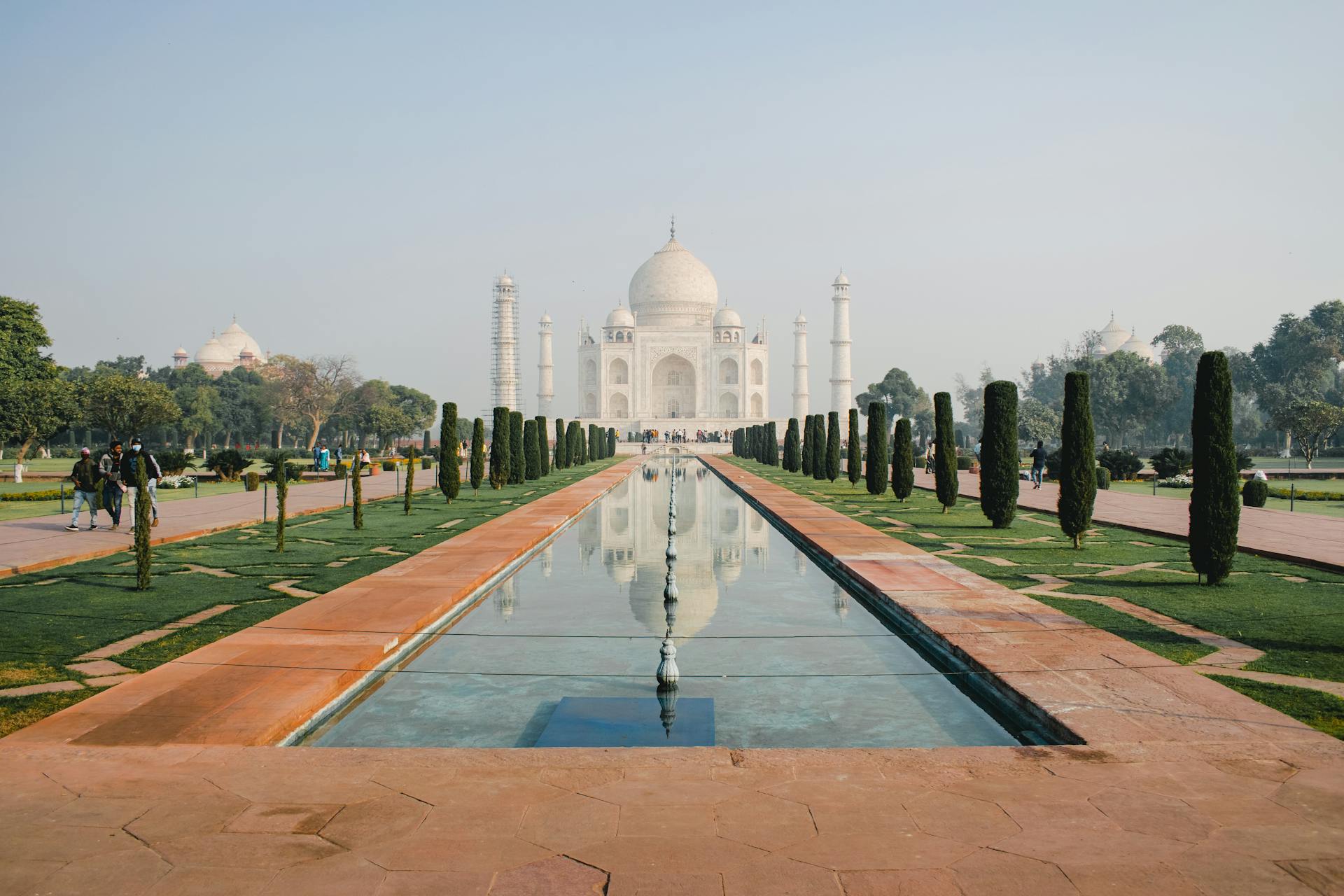 The iconic Taj Mahal reflecting in the water on a sunny day, framed by gardens and trees.