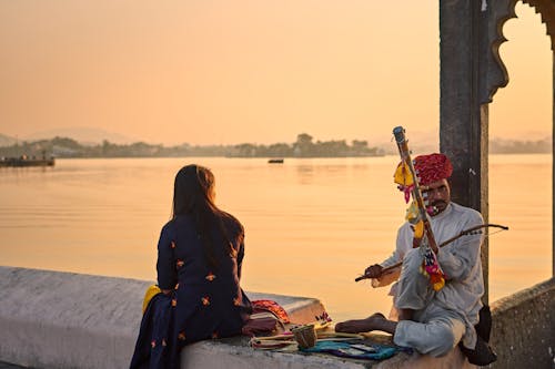 Woman and Man in Traditional Clothing Sitting on Wall on Lakeshore and Playing Musical Instrument