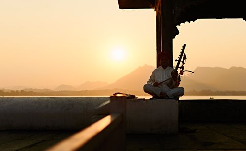 Free A musician playing ravanatha at sunset by a serene lake, dressed in traditional attire. Stock Photo