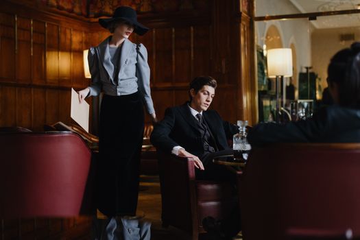 A Woman in Gray Blazer and Black Skirt Standing Near the Man in Black Suit Sitting on the Chair