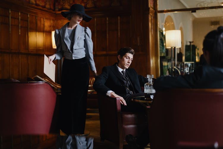 A Woman In Gray Blazer And Black Skirt Standing Near The Man In Black Suit Sitting On The Chair