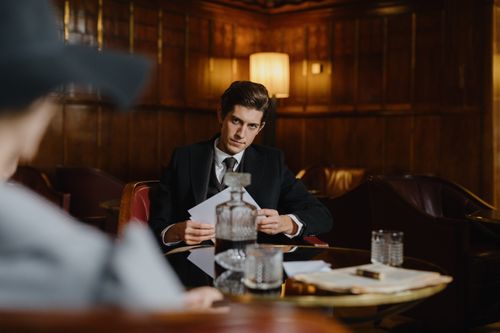 Free A serious man in a suit sitting at a table with papers, evoking a corporate or mobster vibe in an elegant, dimly lit room. Stock Photo
