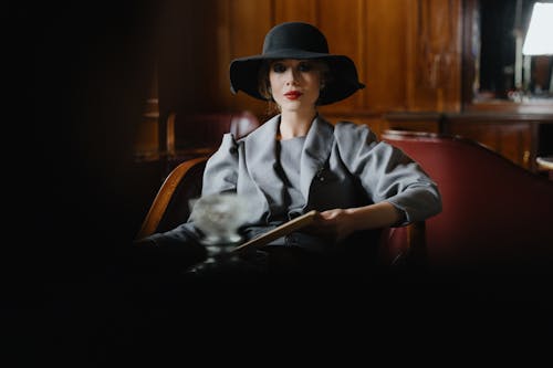 A sophisticated woman in retro style attire with a wide-brimmed hat sitting elegantly indoors.