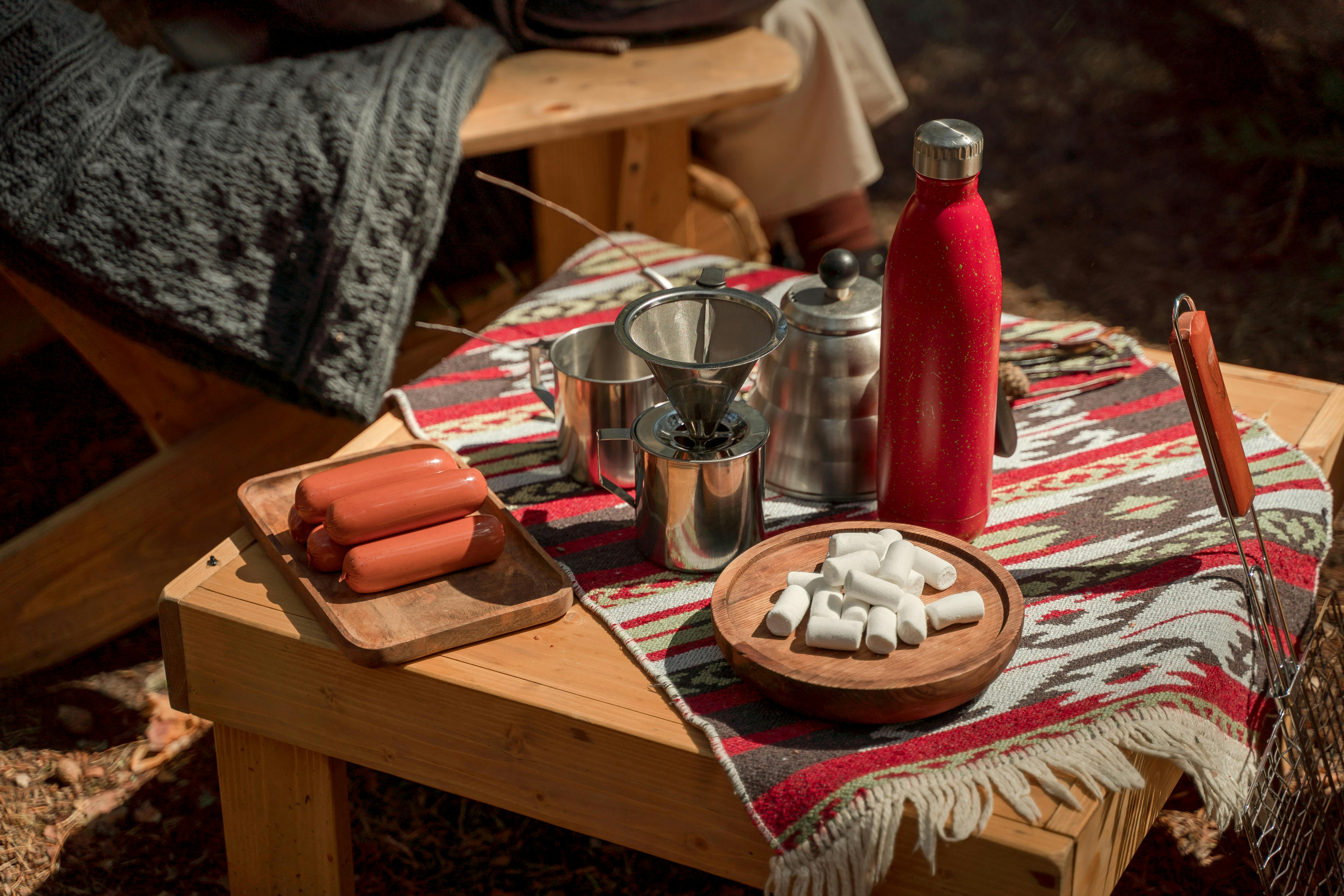 Warm outdoor picnic scene featuring sausages, marshmallows, and coffee on a wooden table.