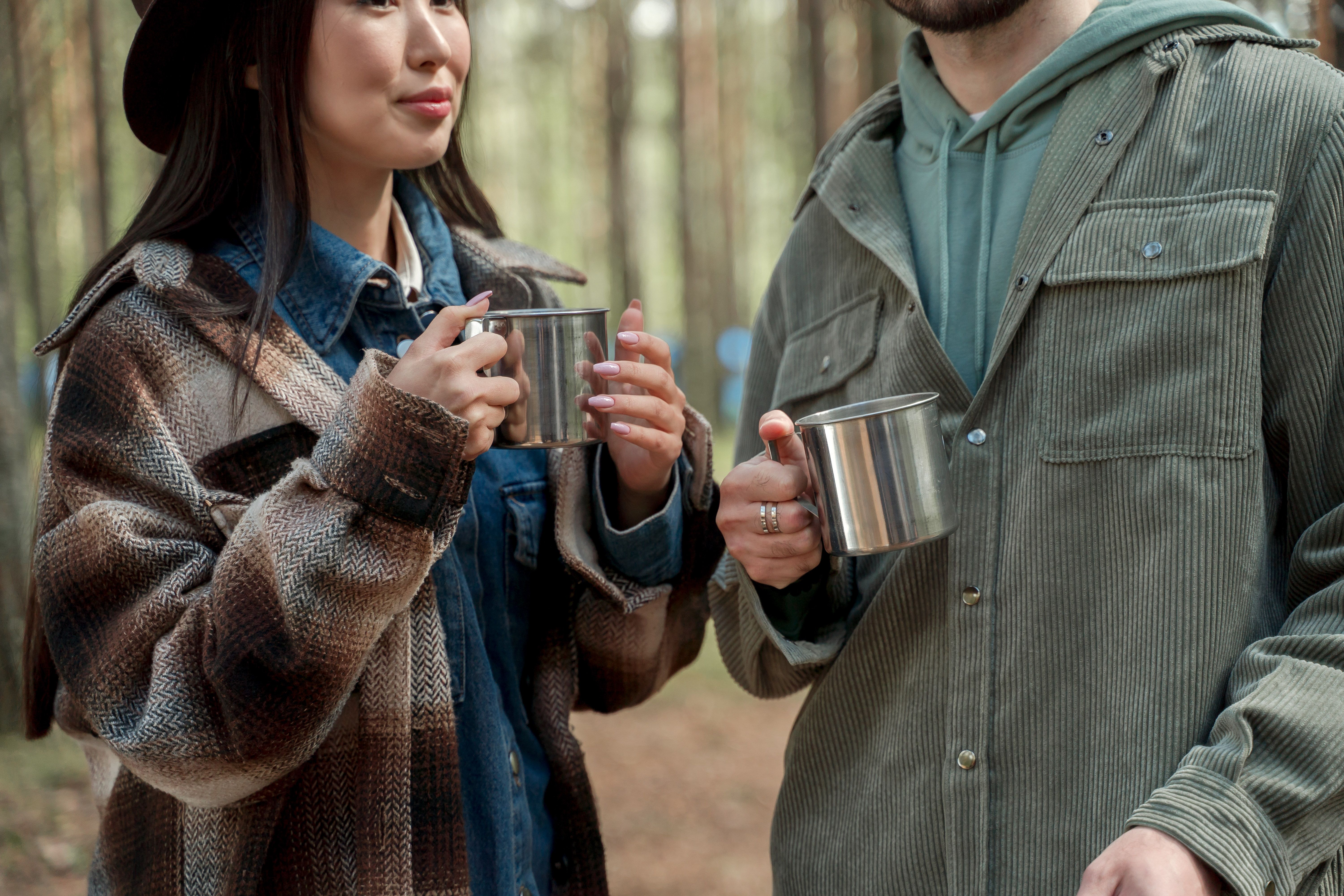 A couple enjoying coffee in stainless mugs during a cozy outdoor excursion in the woods.